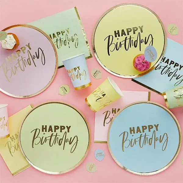 Pastel party setup with scalloped plates, striped cups, party hats, small cakes, and drinks arranged on a pink background